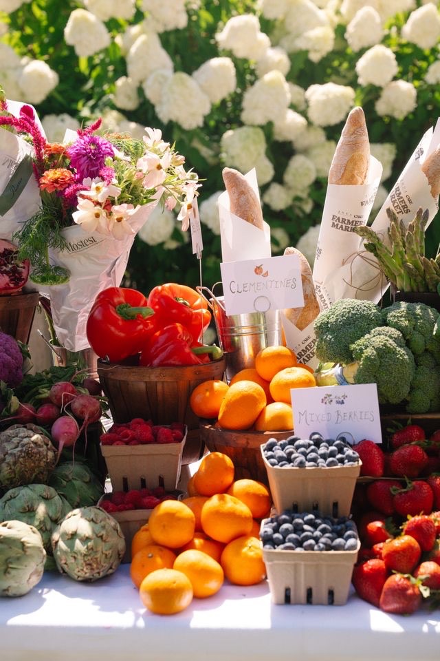 Fresh produce, flowers, artisan bread, and market goods displayed on a beautifully styled outdoor farmer's market table at the Agua by Agua Bendita x CAKE brand activation in the Hamptons.